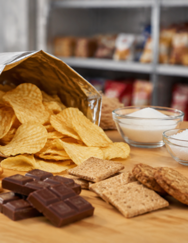 Open bag of potato chips spilling onto a wooden table with crackers, cookies, chocolate, and bowls of sugar and flour nearby.