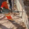 Construction worker in orange reflective pants and gloves uses a jackhammer to break a concrete wall, with rubble and dirt around.