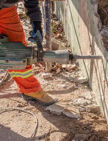 Construction worker in orange reflective pants and gloves uses a jackhammer to break a concrete wall, with rubble and dirt around.