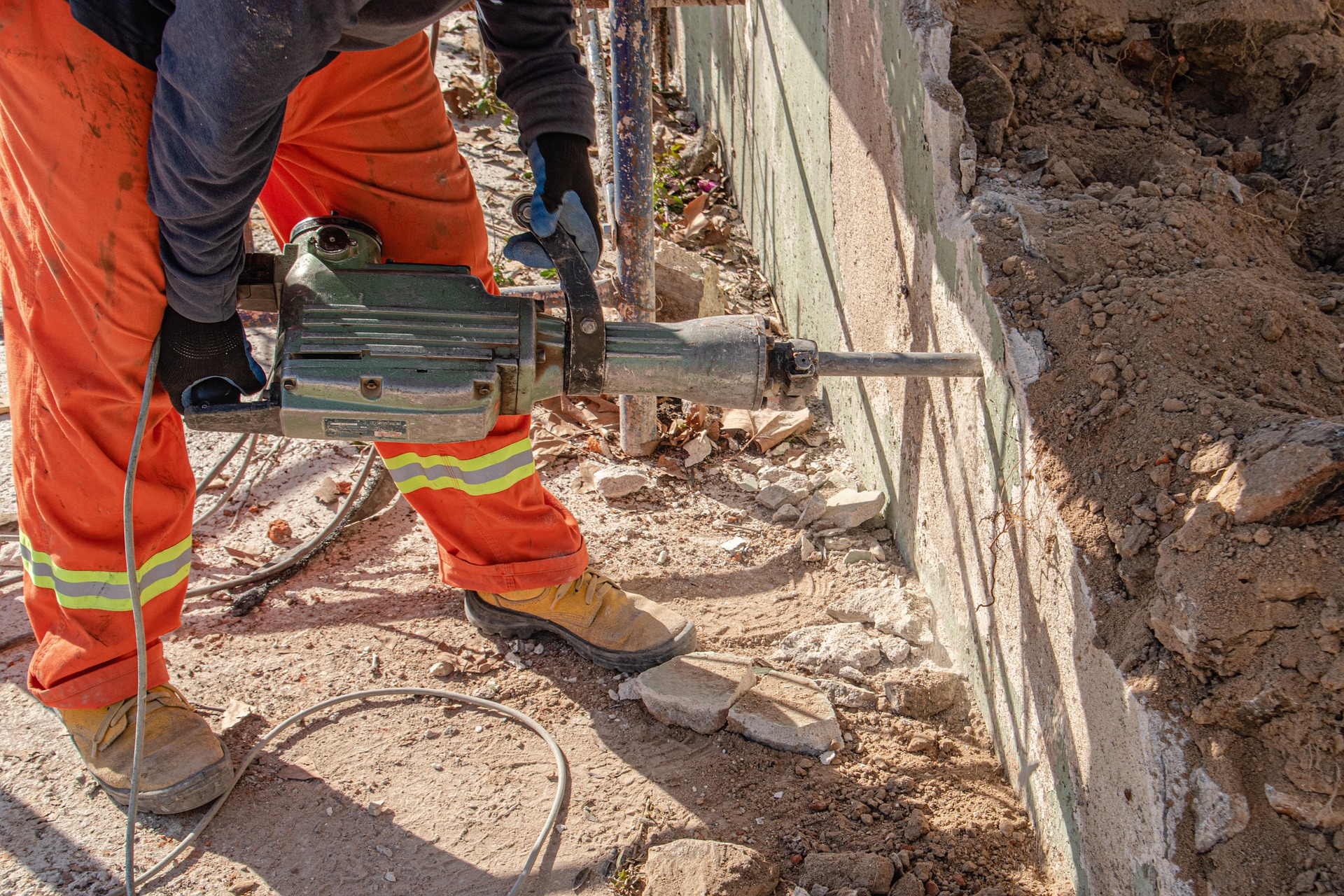Construction worker in orange reflective pants and gloves uses a jackhammer to break a concrete wall, with rubble and dirt around.