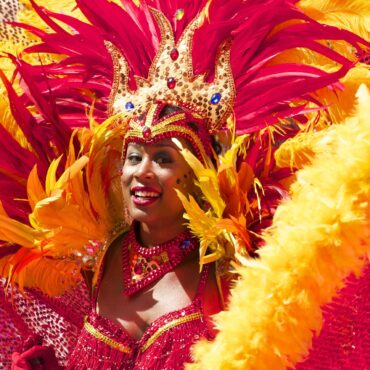 Carnival performer in a red-gold feather headdress with sequined crown, smiling at the camera.