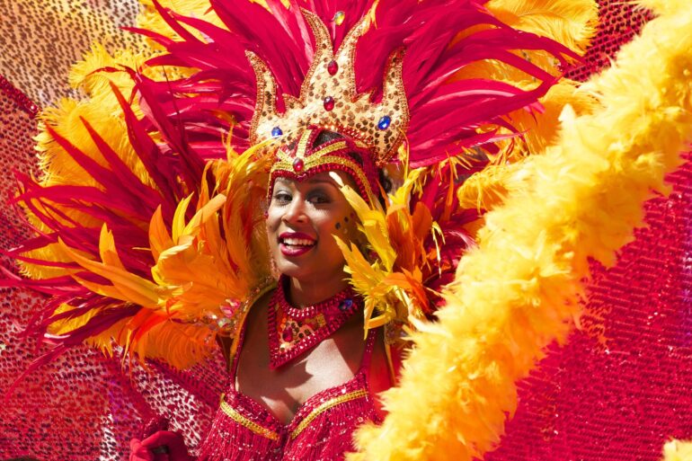 Carnival performer in a red-gold feather headdress with sequined crown, smiling at the camera.