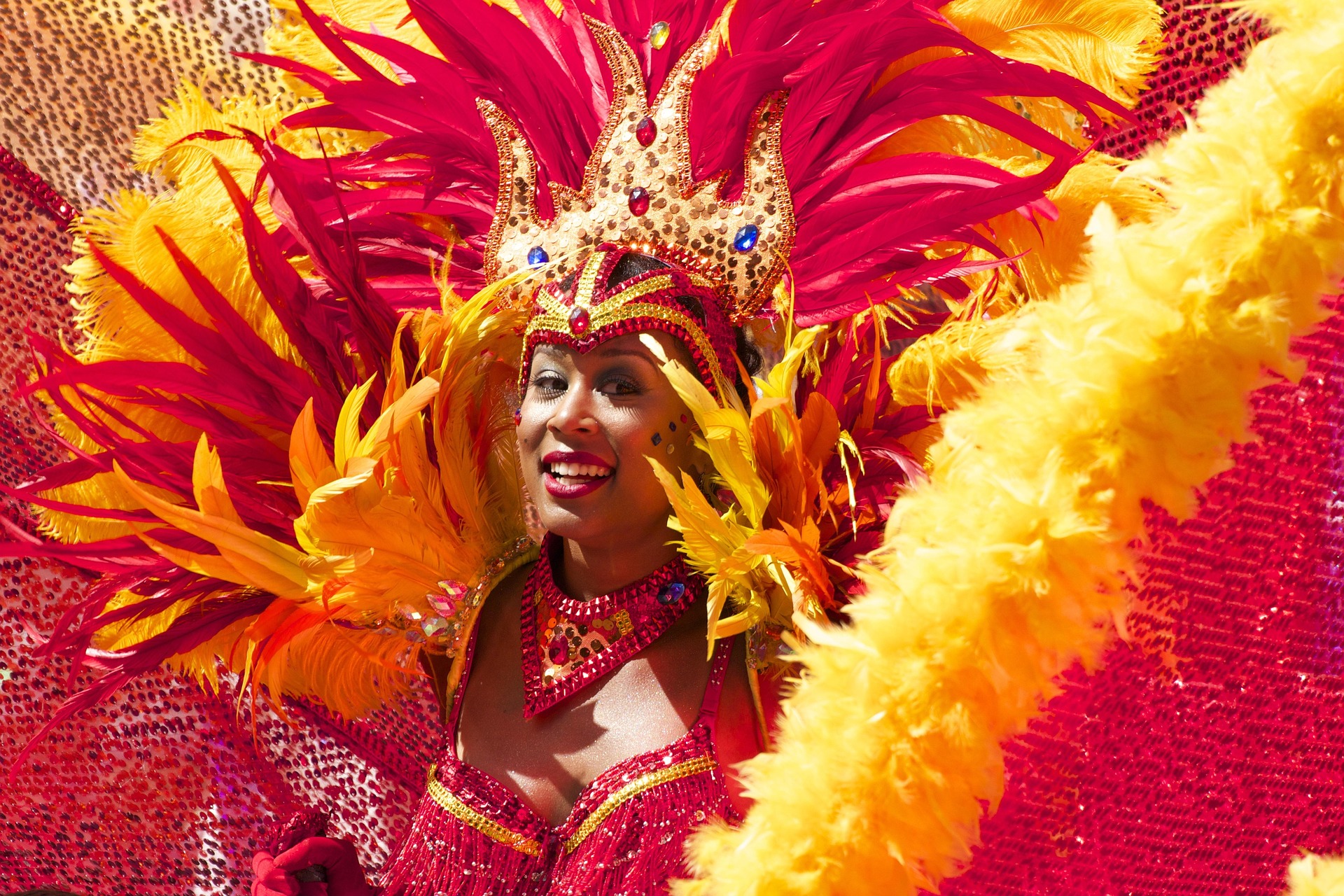 Carnival performer in a red-gold feather headdress with sequined crown, smiling at the camera.