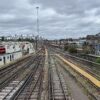 View down a set of parallel train tracks converging toward a distant city skyline under an overcast sky, with station platforms on the sides.