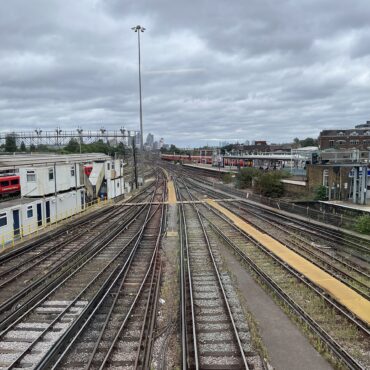 View down a set of parallel train tracks converging toward a distant city skyline under an overcast sky, with station platforms on the sides.