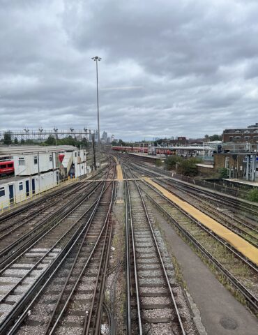 View down a set of parallel train tracks converging toward a distant city skyline under an overcast sky, with station platforms on the sides.