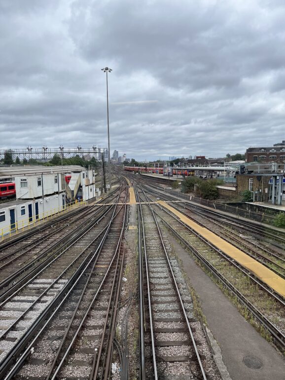 Clapham_Junction_in_2022 - NOTE.radio - Polskie radio w UK | Polskie radio w Londynie View down a set of parallel train tracks converging toward a distant city skyline under an overcast sky, with station platforms on the sides.
