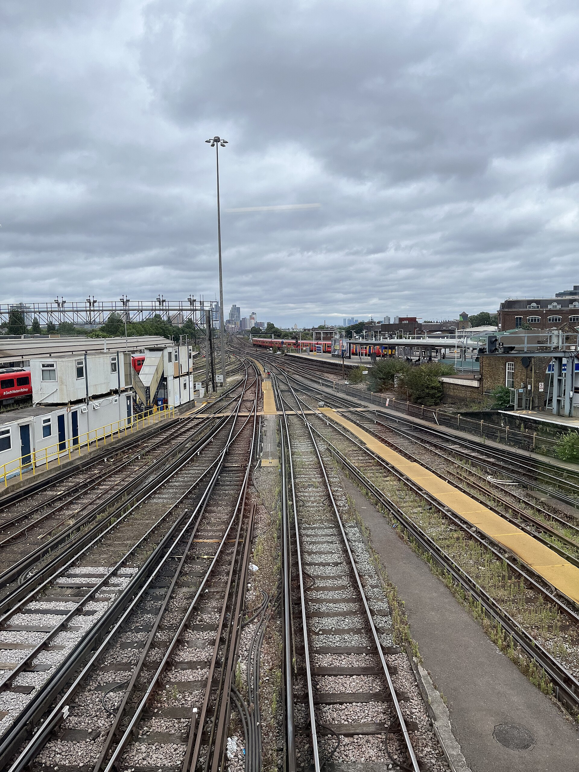 View down a set of parallel train tracks converging toward a distant city skyline under an overcast sky, with station platforms on the sides.