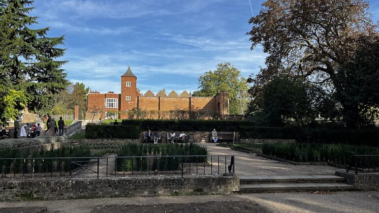Dutch_Garden_and_Holland_House_Holland_Park_-_geographorguk_-_7620545 - NOTE.radio - Polskie radio w UK | Polskie radio w Londynie Brick castle-style wall with a tall tower overlooking a tidy park garden; people stroll and sit along the stone paths on a sunny day.