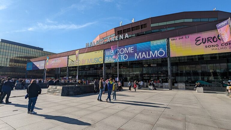 Eurovision_2024_Malmö_Arena_external - NOTE.radio - Polskie radio w UK | Polskie radio w Londynie Exterior view of Malmö Arena with colorful banners reading 'WELCOME BACK TO MALMÖ' and people walking outside on a sunny day.