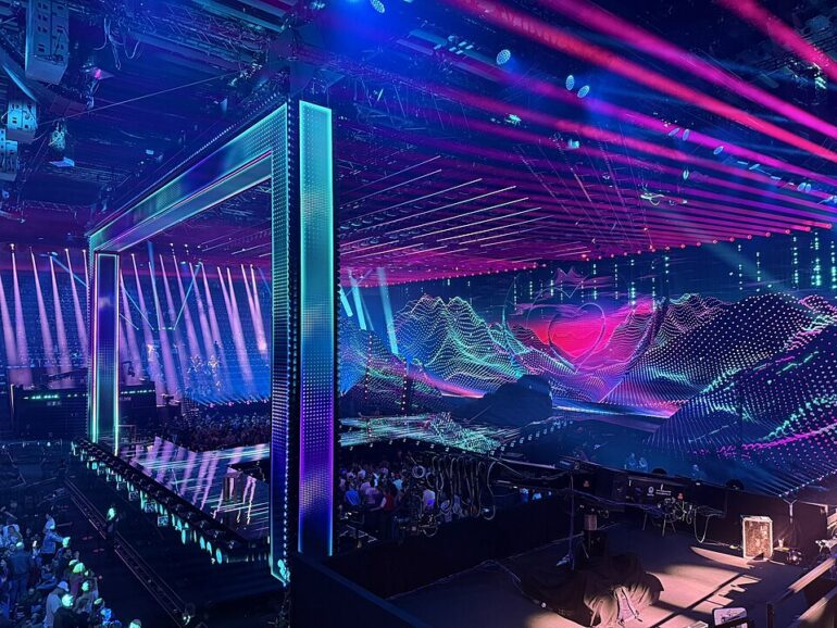 Futuristic nightclub stage with neon frames, purple-blue lasers, and a LED backdrop; crowd seated and watching a performance.