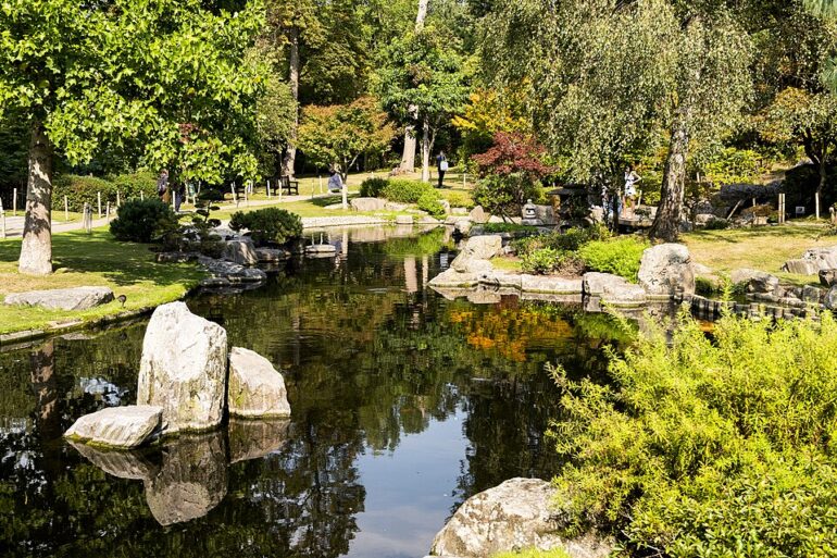 Kyoto_Garden_Holland_Park - NOTE.radio - Polskie radio w UK | Polskie radio w Londynie A tranquil park pond with large rocks, green trees, and a few people walking along the paths nearby, reflecting in the water.