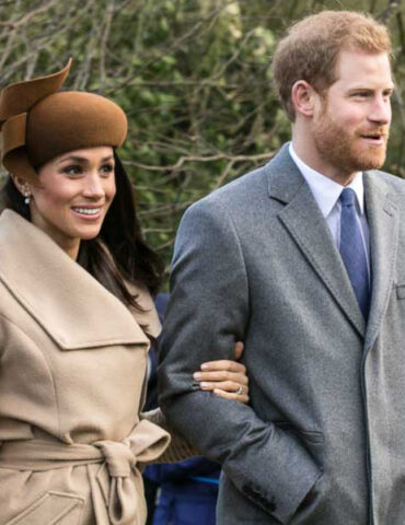 A smiling couple walking outdoors; woman in a beige coat with a brown hat and the man in a gray suit and blue tie walking side by side, arm linked.