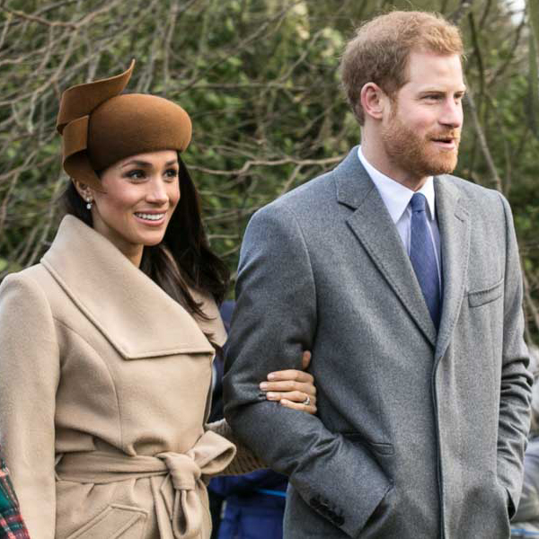 A smiling couple walking outdoors; woman in a beige coat with a brown hat and the man in a gray suit and blue tie walking side by side, arm linked.