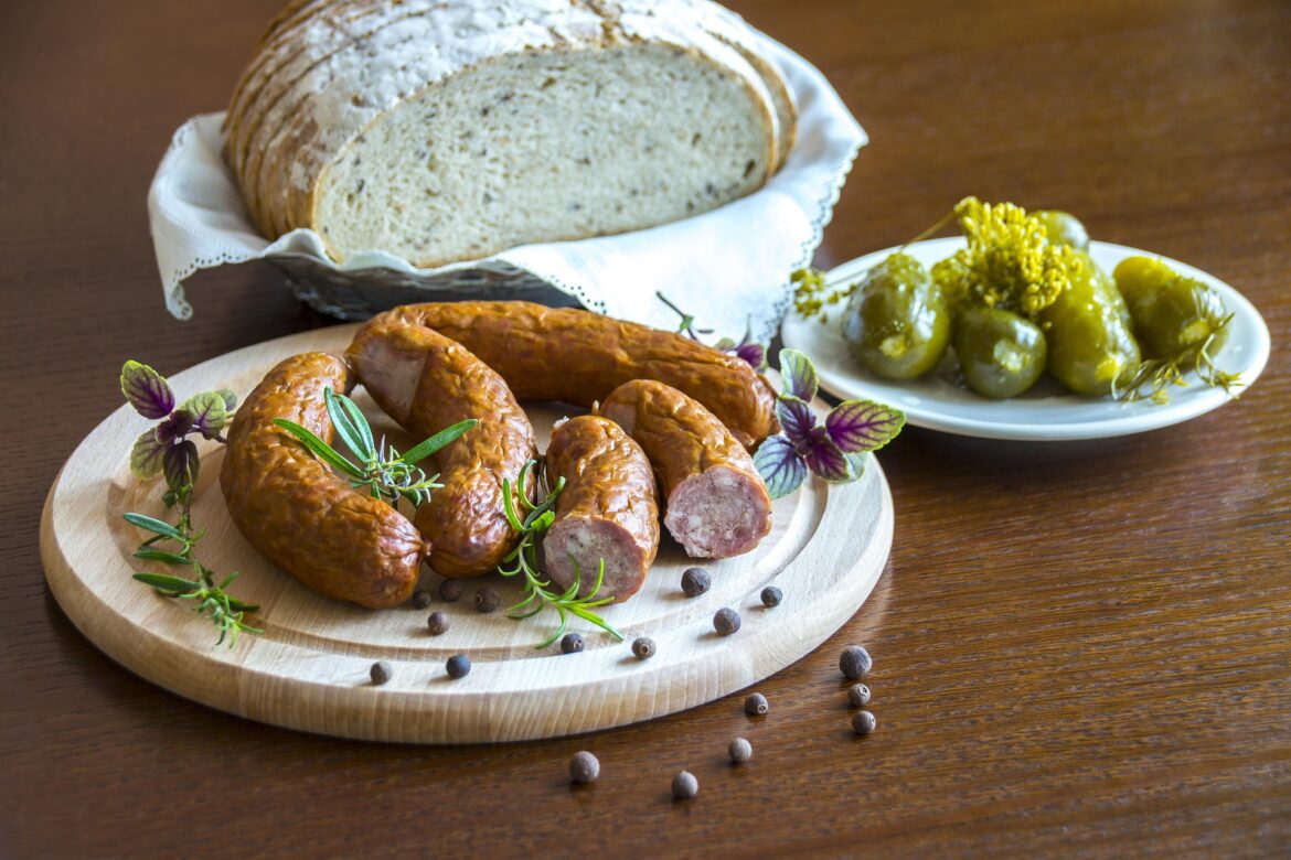 Rustic loaf of bread on a napkin with sausages on a wooden board, garnished with rosemary and peppercorns; pickles on a white plate nearby.