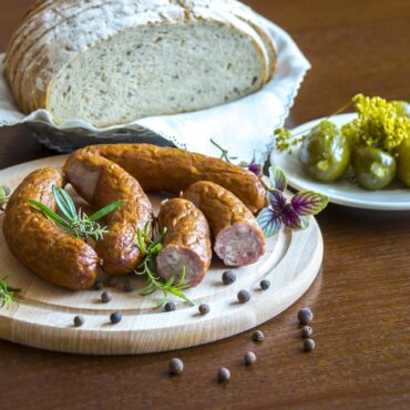 Rustic loaf of bread on a napkin with sausages on a wooden board, garnished with rosemary and peppercorns; pickles on a white plate nearby.