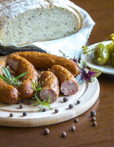 Rustic loaf of bread on a napkin with sausages on a wooden board, garnished with rosemary and peppercorns; pickles on a white plate nearby.
