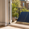 Two solar panels on a stand sit on a sunlit balcony, connected by a cable to an indoor power outlet.