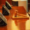 Close-up of a wooden judge's gavel resting on a desk with blurred conference devices in the background, suggesting a legal setting.