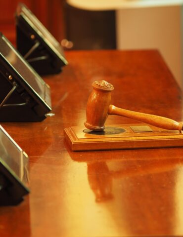 Close-up of a wooden judge's gavel resting on a desk with blurred conference devices in the background, suggesting a legal setting.
