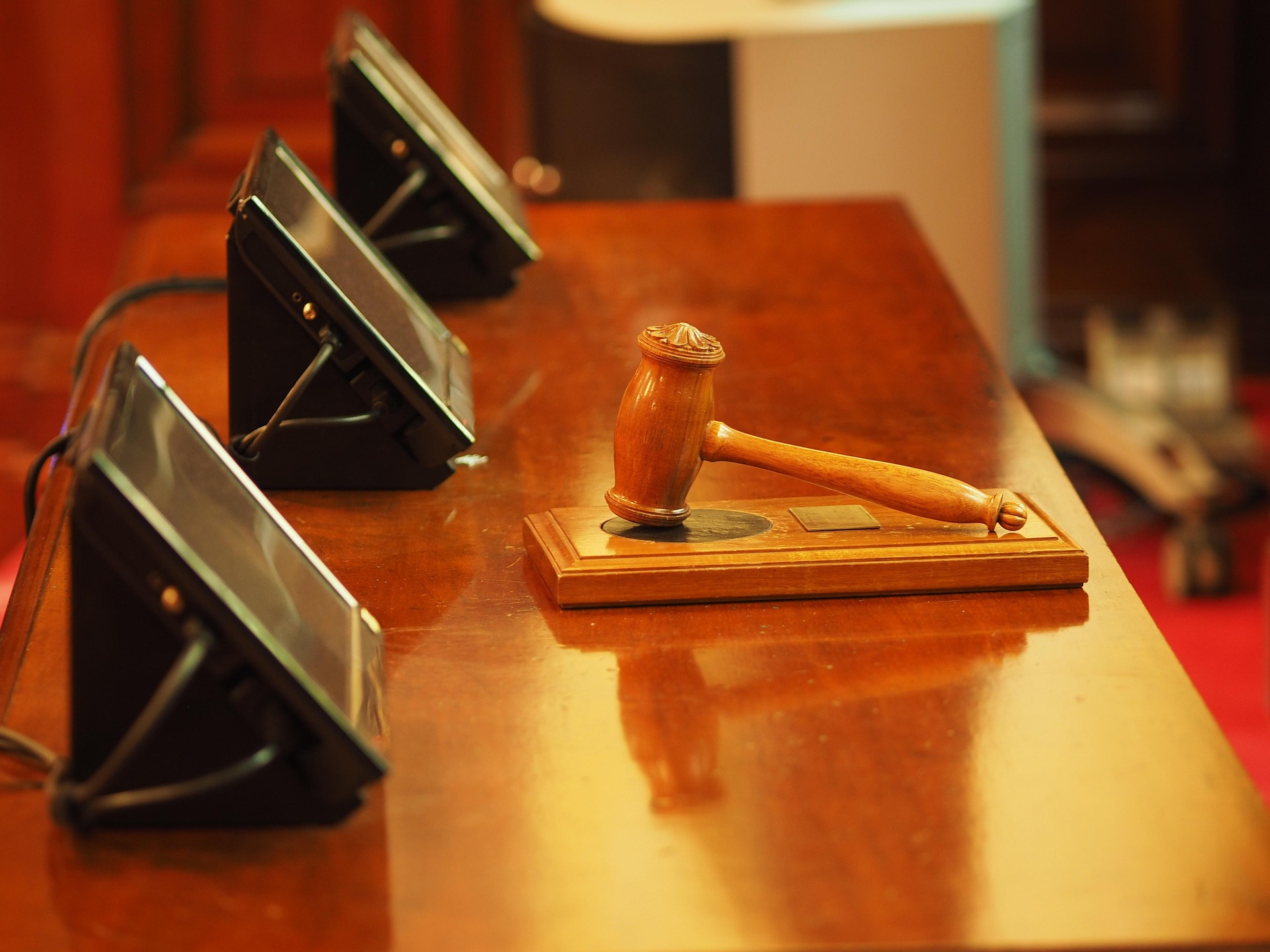 Close-up of a wooden judge's gavel resting on a desk with blurred conference devices in the background, suggesting a legal setting.