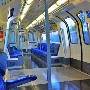 Inside a passenger train car with blue patterned seats along a narrow aisle and overhead lighting.
