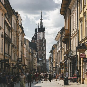 Bustling narrow street lined with historic buildings toward a tall church tower in the distance; people walk, shops on both sides.