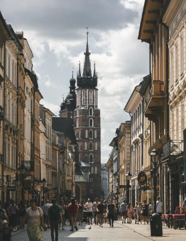 Bustling narrow street lined with historic buildings toward a tall church tower in the distance; people walk, shops on both sides.