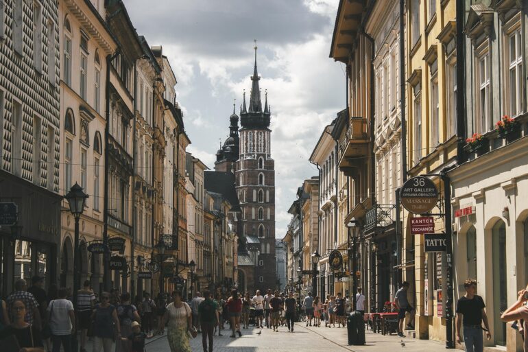 dimaliss-street-4472321_1920 - NOTE.radio - Polskie radio w UK | Polskie radio w Londynie Bustling narrow street lined with historic buildings toward a tall church tower in the distance; people walk, shops on both sides.
