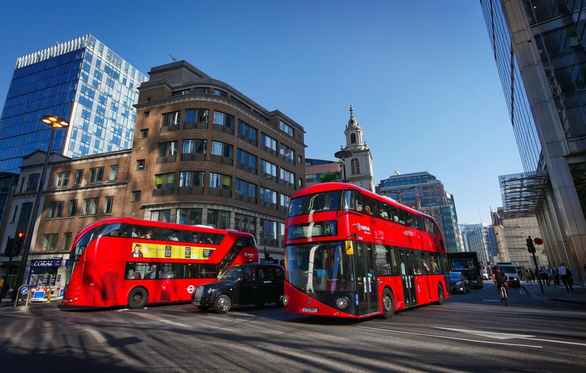 Two red double-decker buses on a busy London street with modern glass and brick buildings under a clear blue sky.