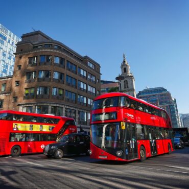 Two red double-decker buses on a busy London street with modern glass and brick buildings under a clear blue sky.
