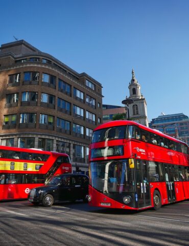 Two red double-decker buses on a busy London street with modern glass and brick buildings under a clear blue sky.