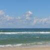 Wide view of a calm ocean meeting a sandy beach under a bright blue sky with scattered clouds, gentle waves rolling in.
