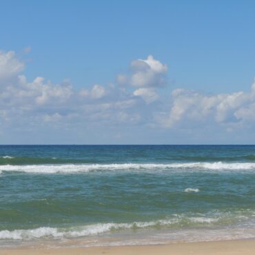 Wide view of a calm ocean meeting a sandy beach under a bright blue sky with scattered clouds, gentle waves rolling in.