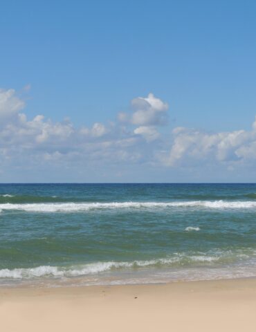 Wide view of a calm ocean meeting a sandy beach under a bright blue sky with scattered clouds, gentle waves rolling in.