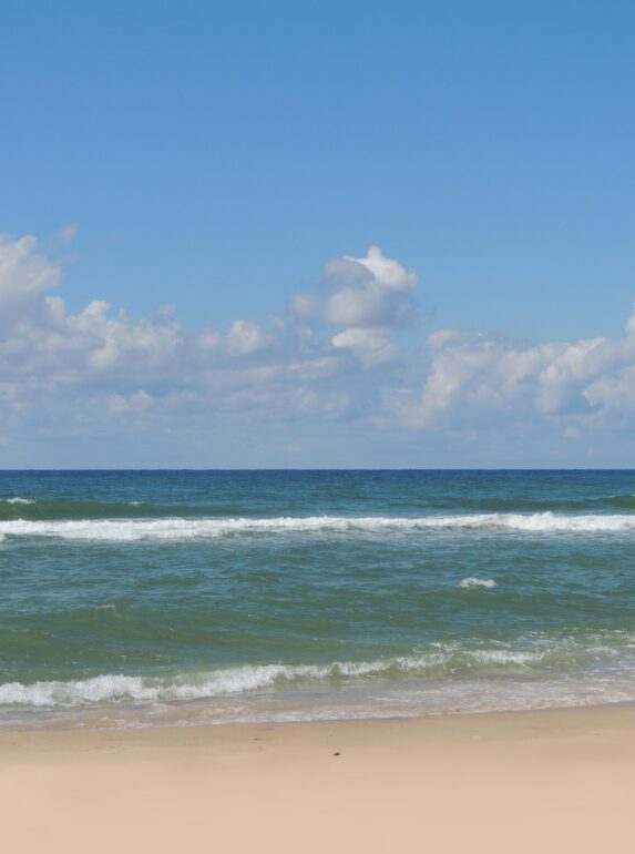 Wide view of a calm ocean meeting a sandy beach under a bright blue sky with scattered clouds, gentle waves rolling in.