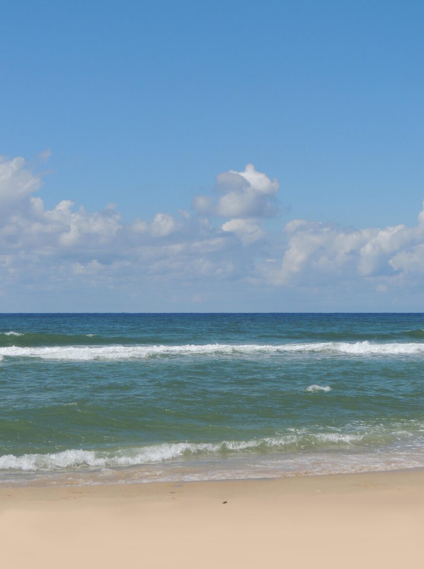 Wide view of a calm ocean meeting a sandy beach under a bright blue sky with scattered clouds, gentle waves rolling in.