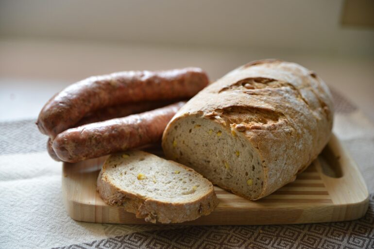 polskie - NOTE.radio - Polskie radio w UK | Polskie radio w Londynie Rustic loaf of bread on a wooden cutting board with a slice cut off, and raw sausages in the background.