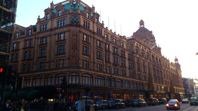 Harrods - NOTE.radio - Polskie radio w UK | Polskie radio w Londynie Harrods department store façade lit with festive string lights on a busy London street at dusk.