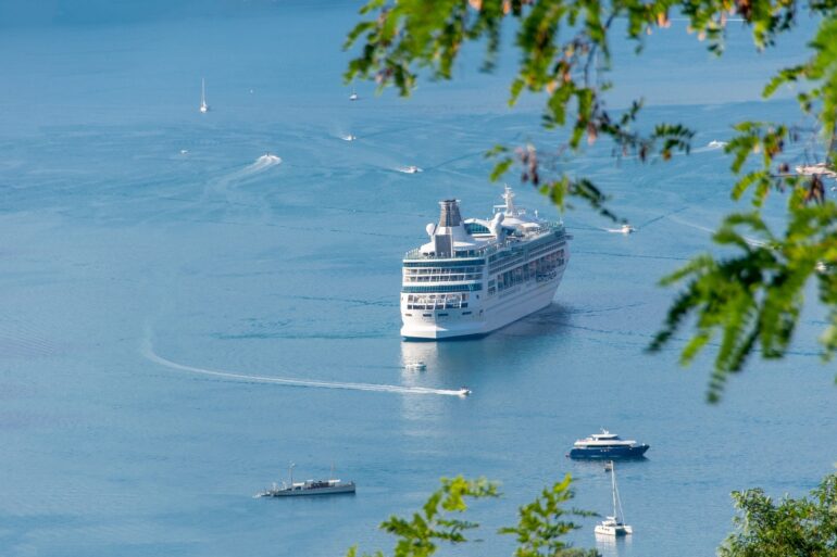 PROM - NOTE.radio - Polskie radio w UK | Polskie radio w Londynie Large white cruise ship cruising on calm blue water, with several small boats nearby and green leaves framing the scene.