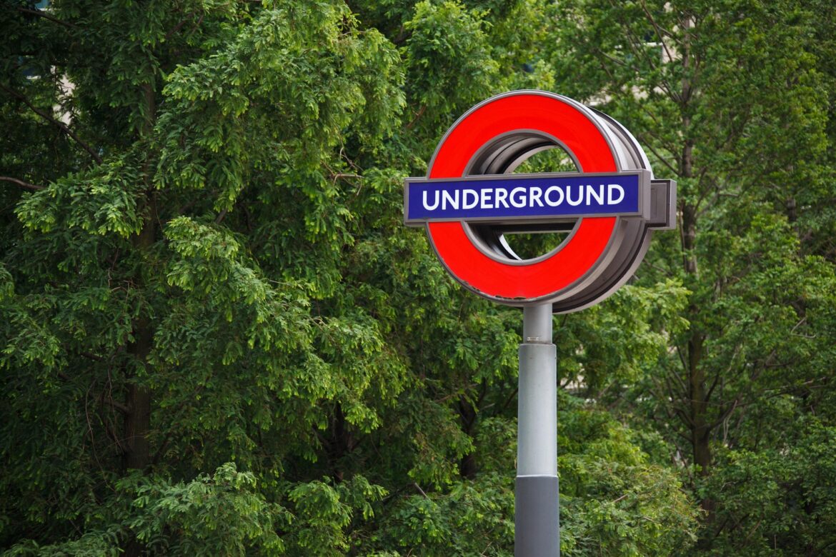 London Underground roundel sign (red circle with blue bar) against green trees in background on a pole.