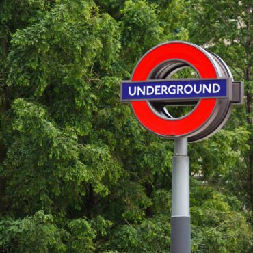 London Underground roundel sign (red circle with blue bar) against green trees in background on a pole.