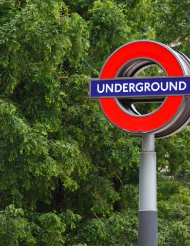 London Underground roundel sign (red circle with blue bar) against green trees in background on a pole.