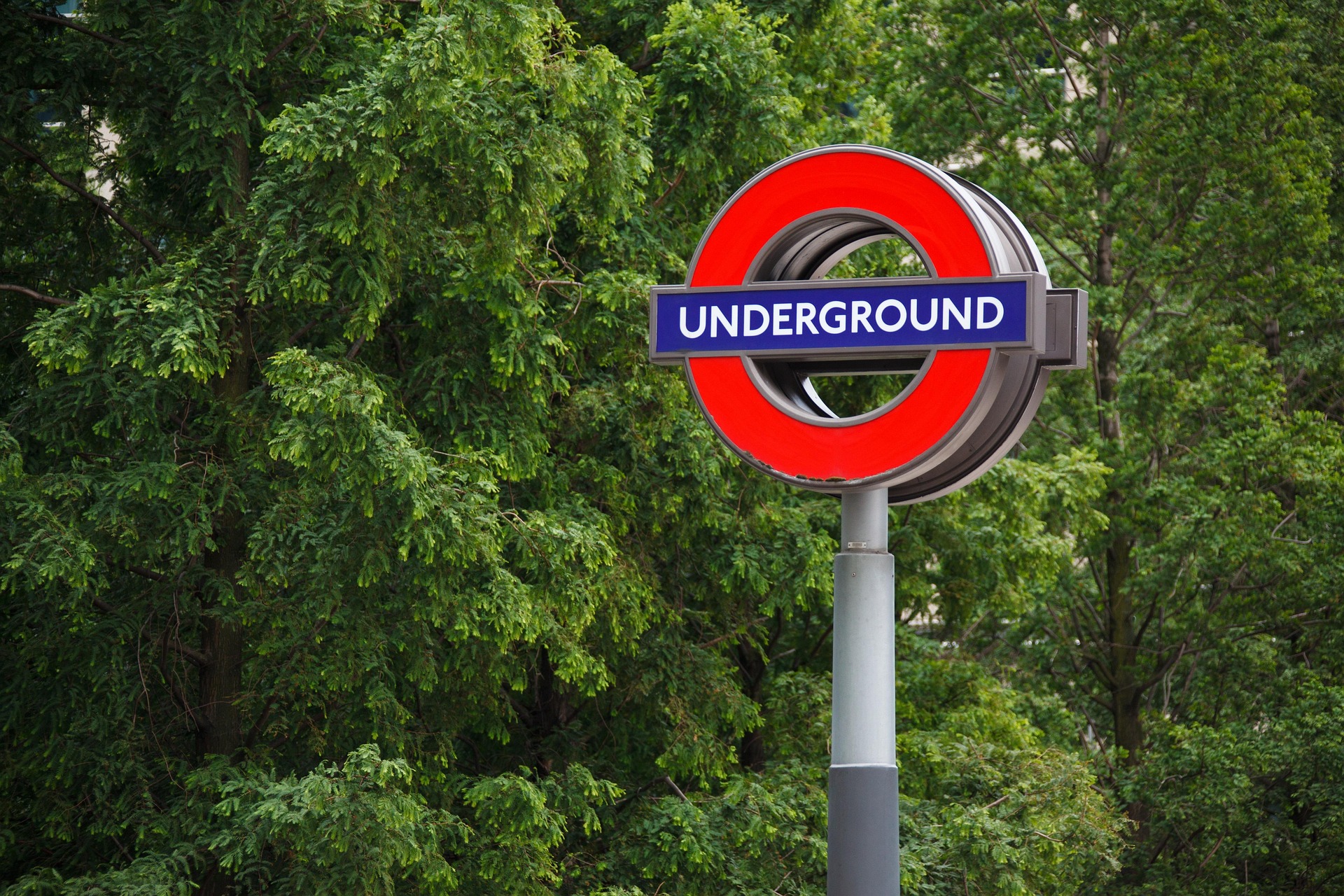 London Underground roundel sign (red circle with blue bar) against green trees in background on a pole.