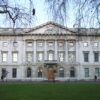 Neoclassical stone building with columns and a triangular pediment, viewed from a park with bare tree branches.