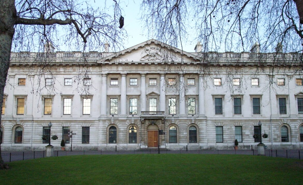 Neoclassical stone building with columns and a triangular pediment, viewed from a park with bare tree branches.