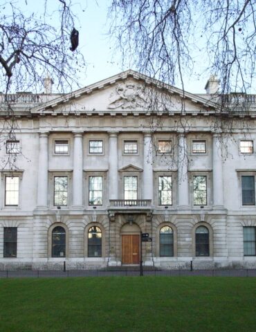 Neoclassical stone building with columns and a triangular pediment, viewed from a park with bare tree branches.
