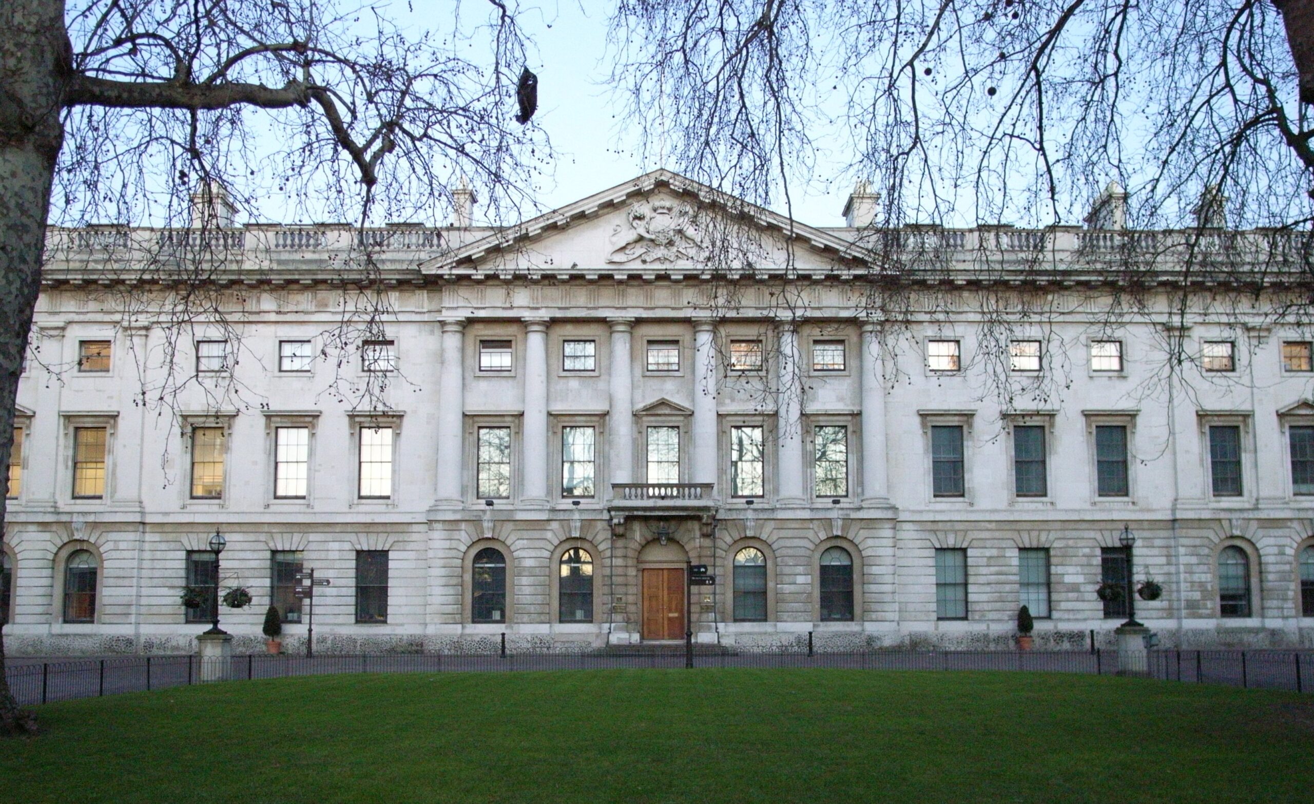 Neoclassical stone building with columns and a triangular pediment, viewed from a park with bare tree branches.
