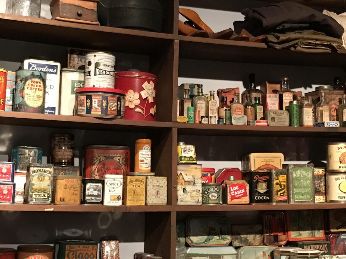 Shelf of vintage metal tins and small glass bottles on dark wooden shelving, resembling an antique shop display with folded items on top.