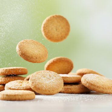 Golden round cookies piled and scattered on a white surface, fine sugar dust floating in the air.